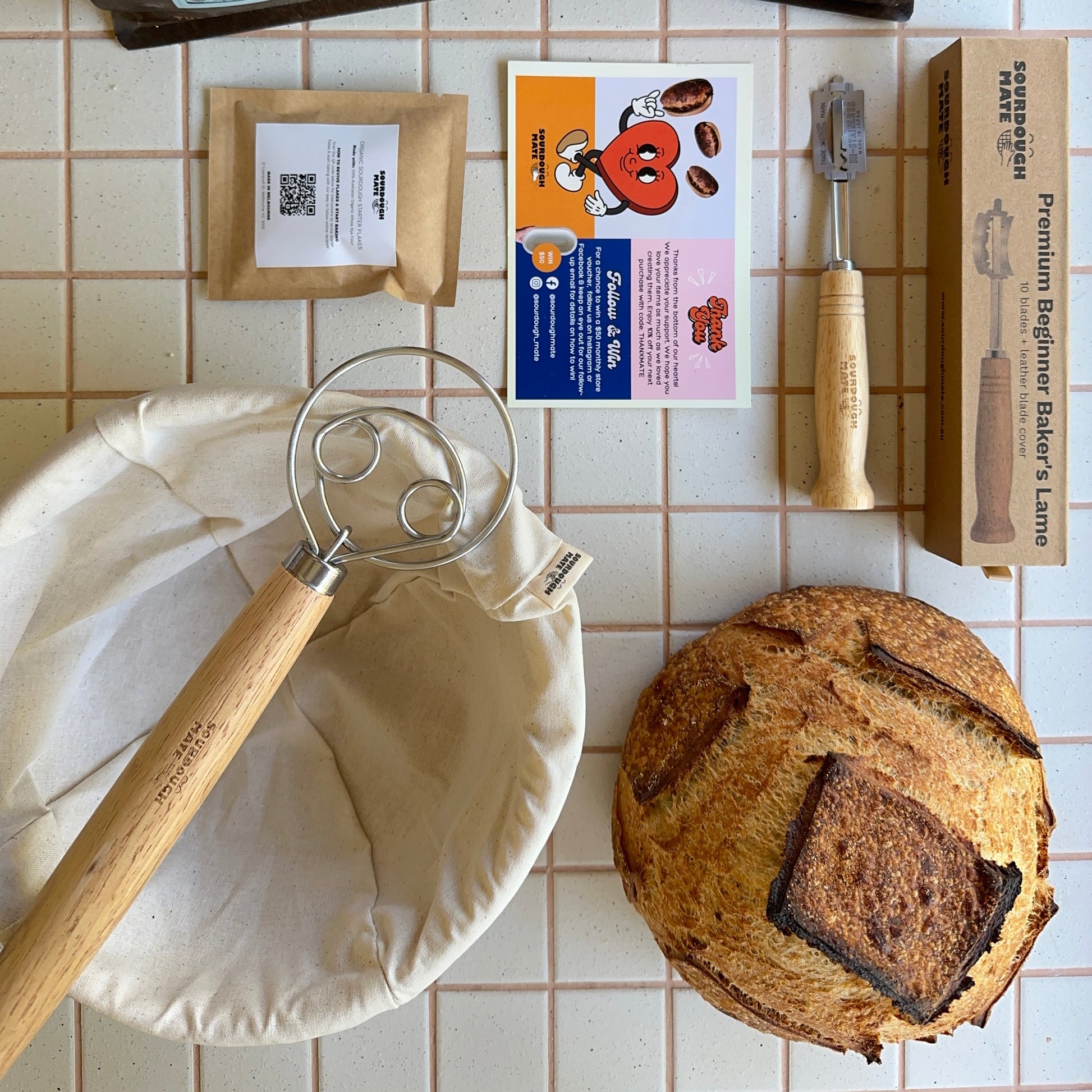 A variety of items laid out on a tiled surface, including a bag of organic wheat flour, a silicon oven mat, a reusable organic cotton bread bag, a wooden dough cutter, and a freshly baked sourdough loaf.