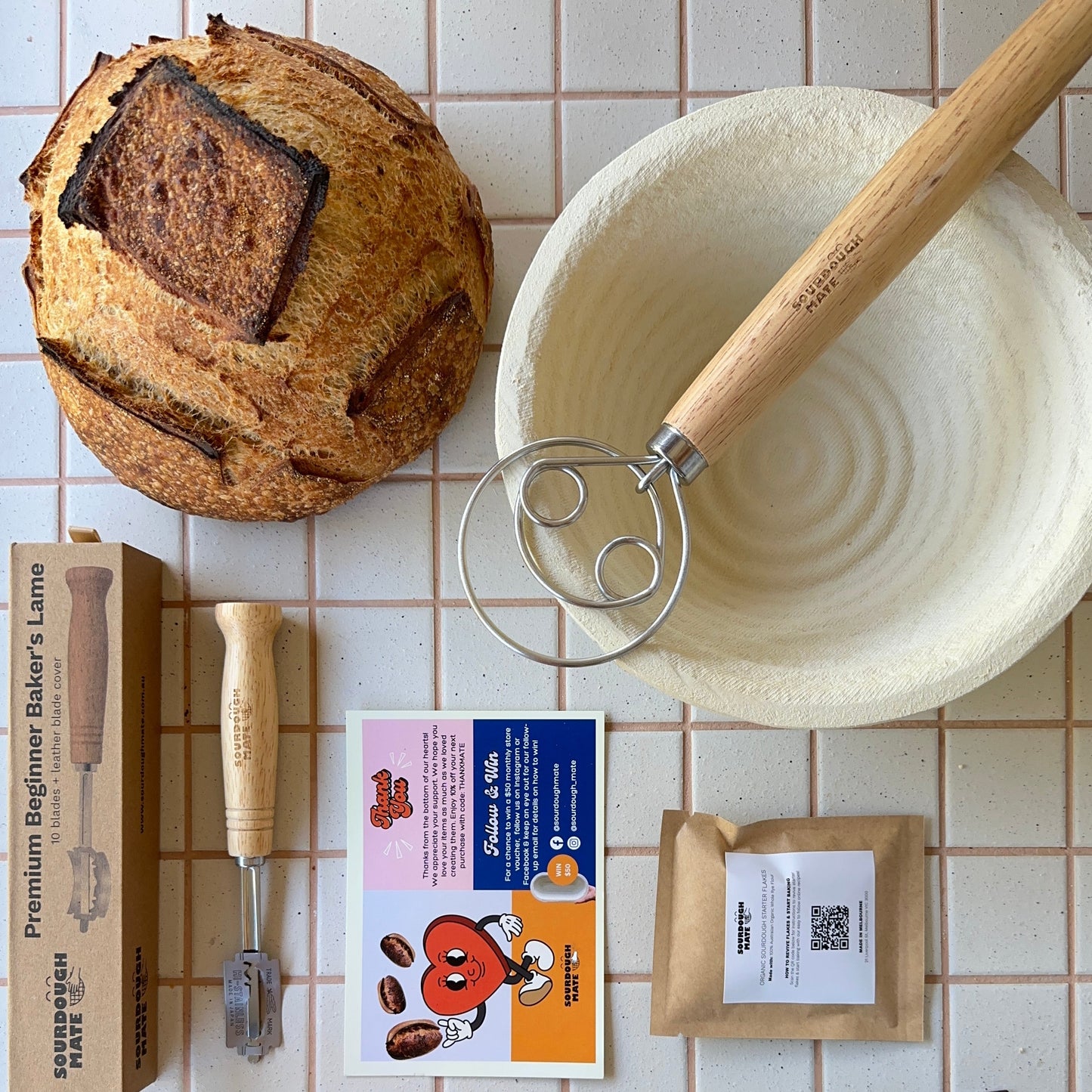 A variety of items laid out on a tiled surface, including a bag of organic wheat flour, a silicon oven mat, a reusable organic cotton bread bag, a wooden dough cutter, and a freshly baked sourdough loaf.