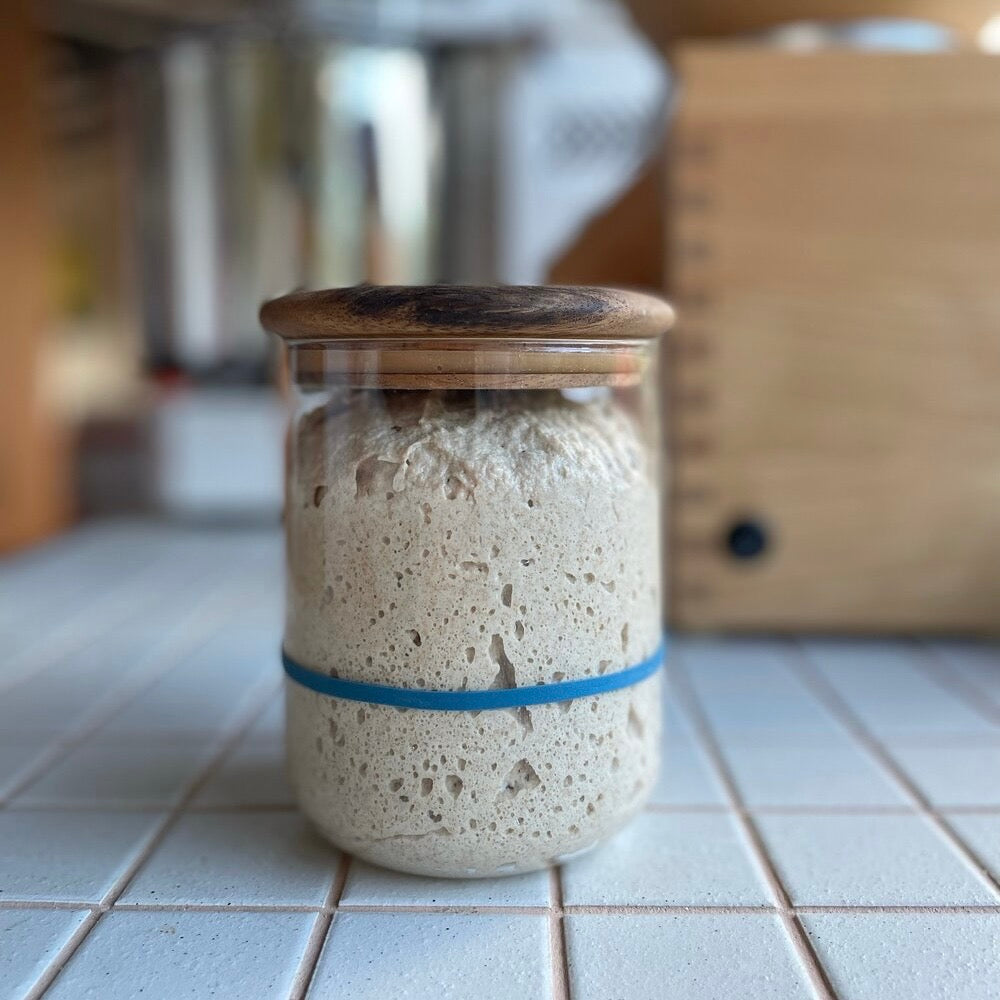 Glass jar with wooden lid containing a dough-like substance on a tiled kitchen floor.