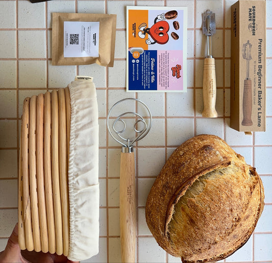 Baking supplies including bags of flour, a loaf of bread, starter, proving basket, lame and a scraper on a tiled surface.