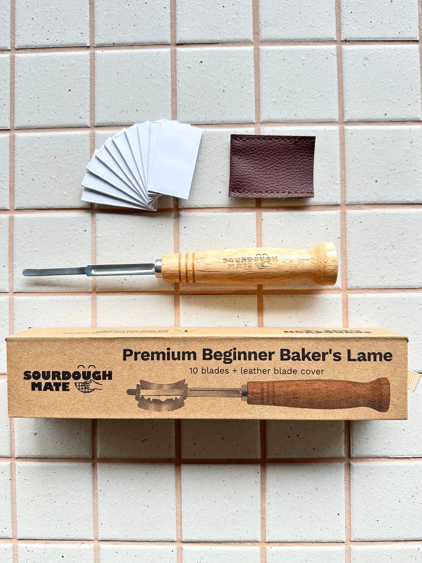 Sourdough Mate Premium Beginner Baker's Lame tool set laid out on a beige tiled background, including a wooden-handled tool, branded cardboard box, 10 replacement blades, and a brown leather blade cover.
