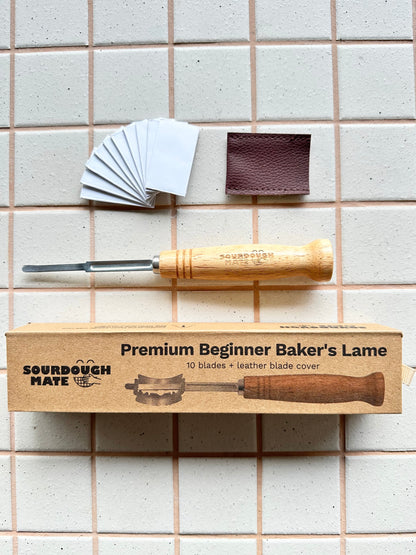 Sourdough Mate Premium Beginner Baker's Lame tool set laid out on a beige tiled background, including a wooden-handled tool, branded cardboard box, 10 replacement blades, and a brown leather blade cover.