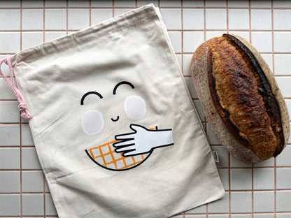 A cream-colored canvas dust bag featuring a minimalist graphic of a hand holding a waffle, styled on a beige tiled surface next to a rustic loaf of bread.