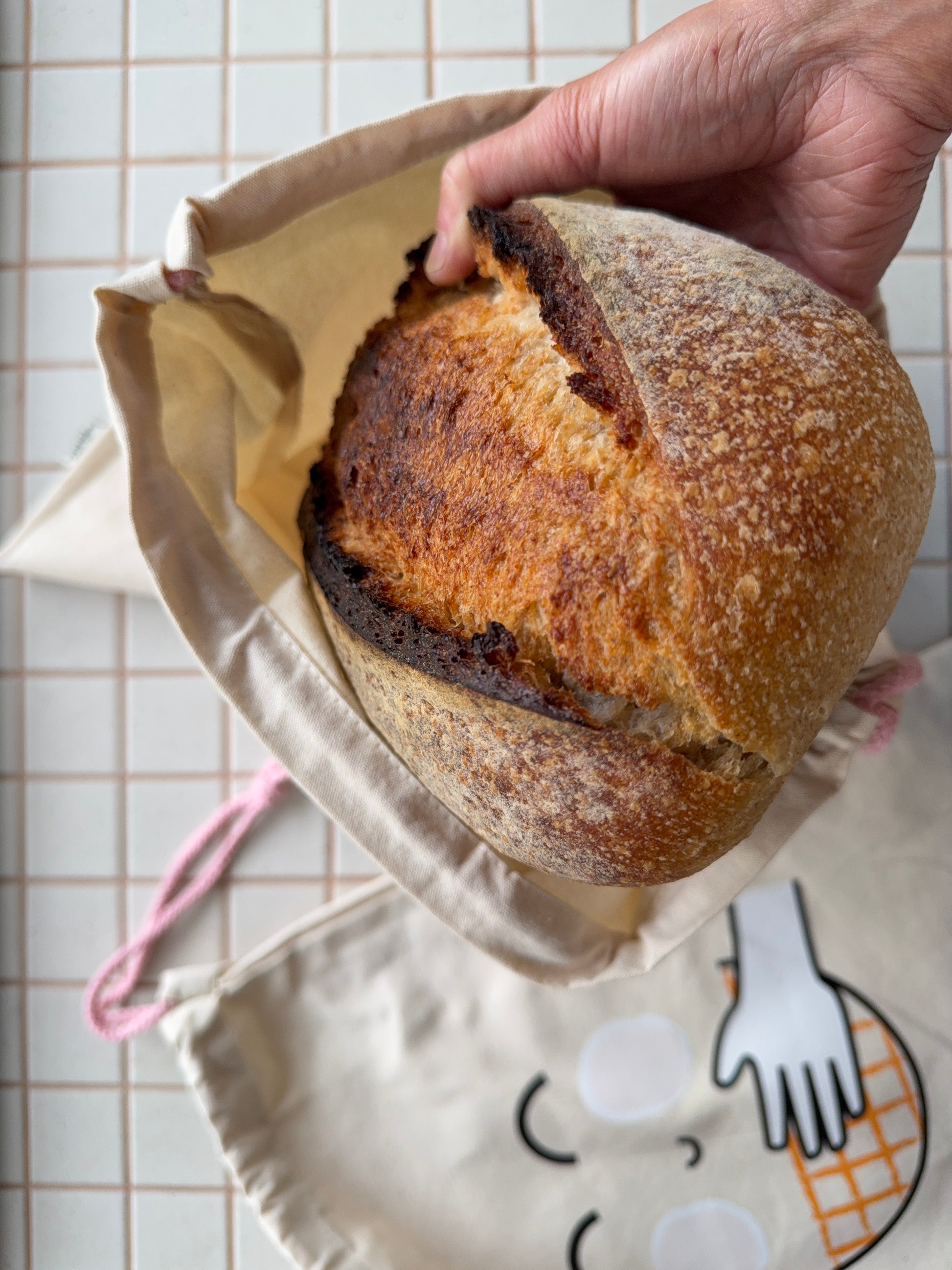 A hand holding a freshly baked sourdough bread loaf inside a beige canvas bag, with another canvas bag featuring a cartoon face and hand graphic lying on a white tiled surface in the background.