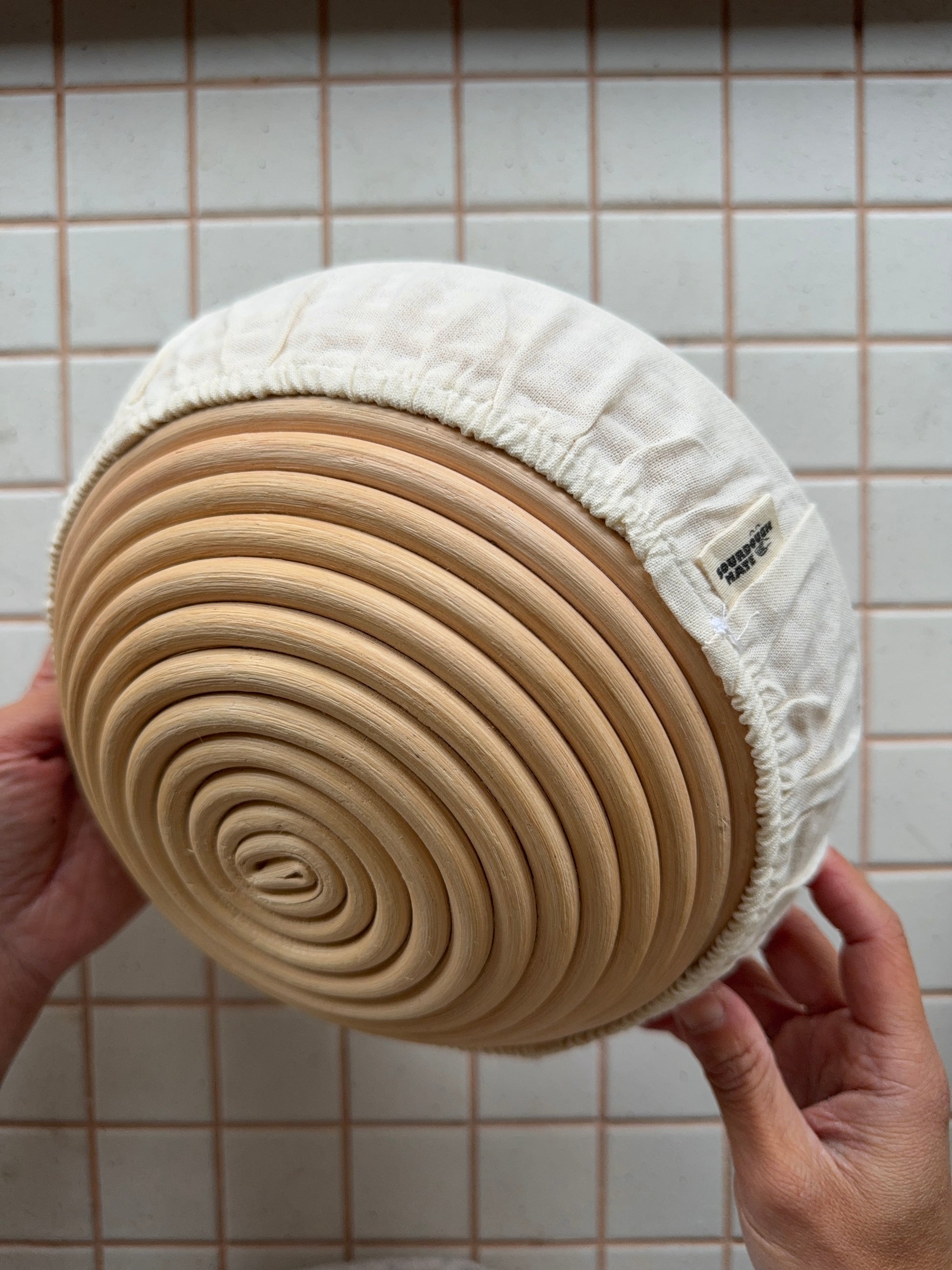 Small Round bread proofing basket with a natural linen cover held by hands against a tiled wall background