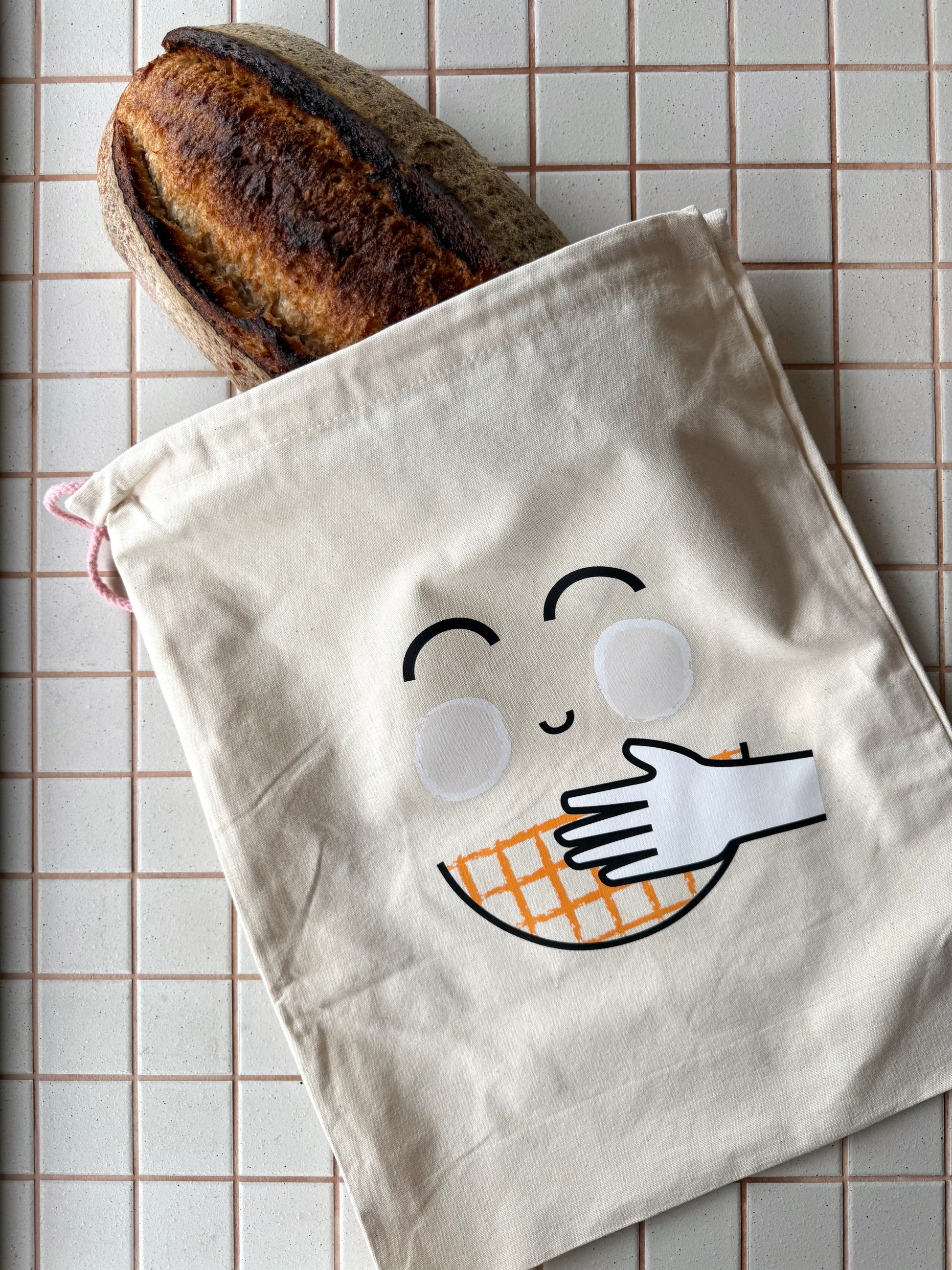 A cream-colored cotton drawstring bag with a cute embroidered face and hand holding an orange-and-white checkered waffle, shown with a loaf of bread peeking out from the top on a beige tiled background.