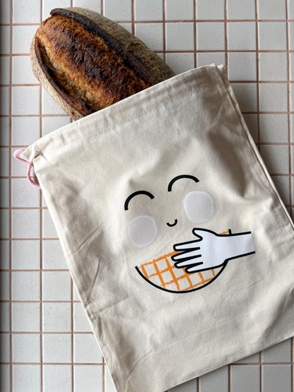A cream-colored cotton drawstring bag with a cute embroidered face and hand holding an orange-and-white checkered waffle, shown with a loaf of bread peeking out from the top on a beige tiled background.