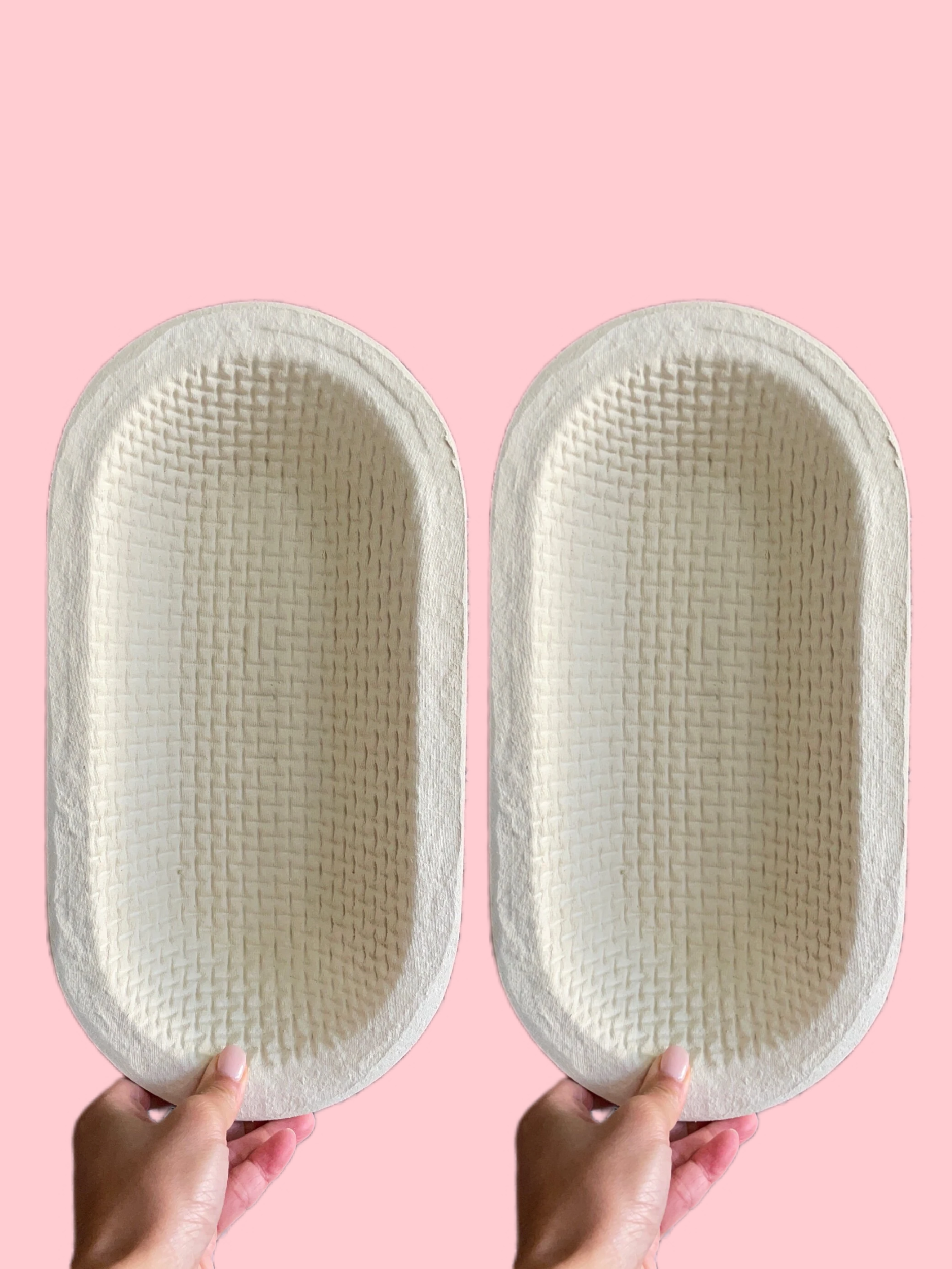 Two oval-shaped beige woven bath mats held up by hands against a solid pink background.