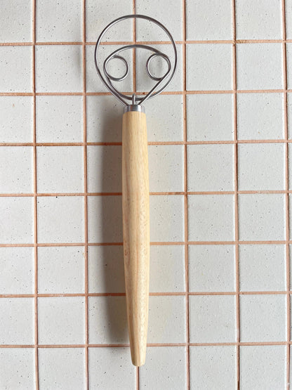A kitchen utensil with a light natural wood handle and a silver metal loop head, shown against a white tiled background.