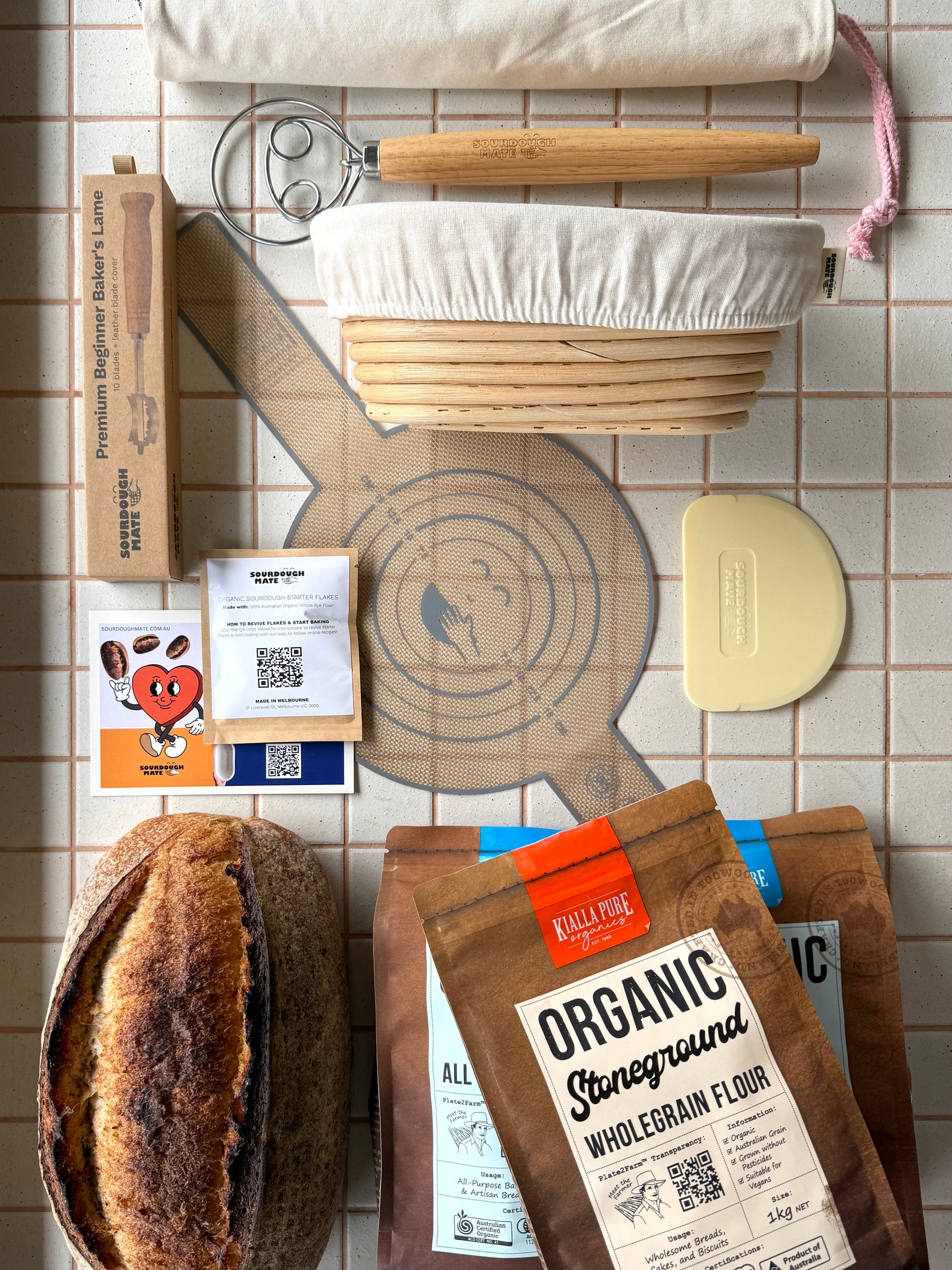 A styled flat-lay of baking tools and ingredients on a beige tiled surface, including a danish dough whisk, cloth-lined banneton, 3kg organic flour, silicon bread sling, organic cotton bread bag, bowl scraper, lame, starter flakes. 