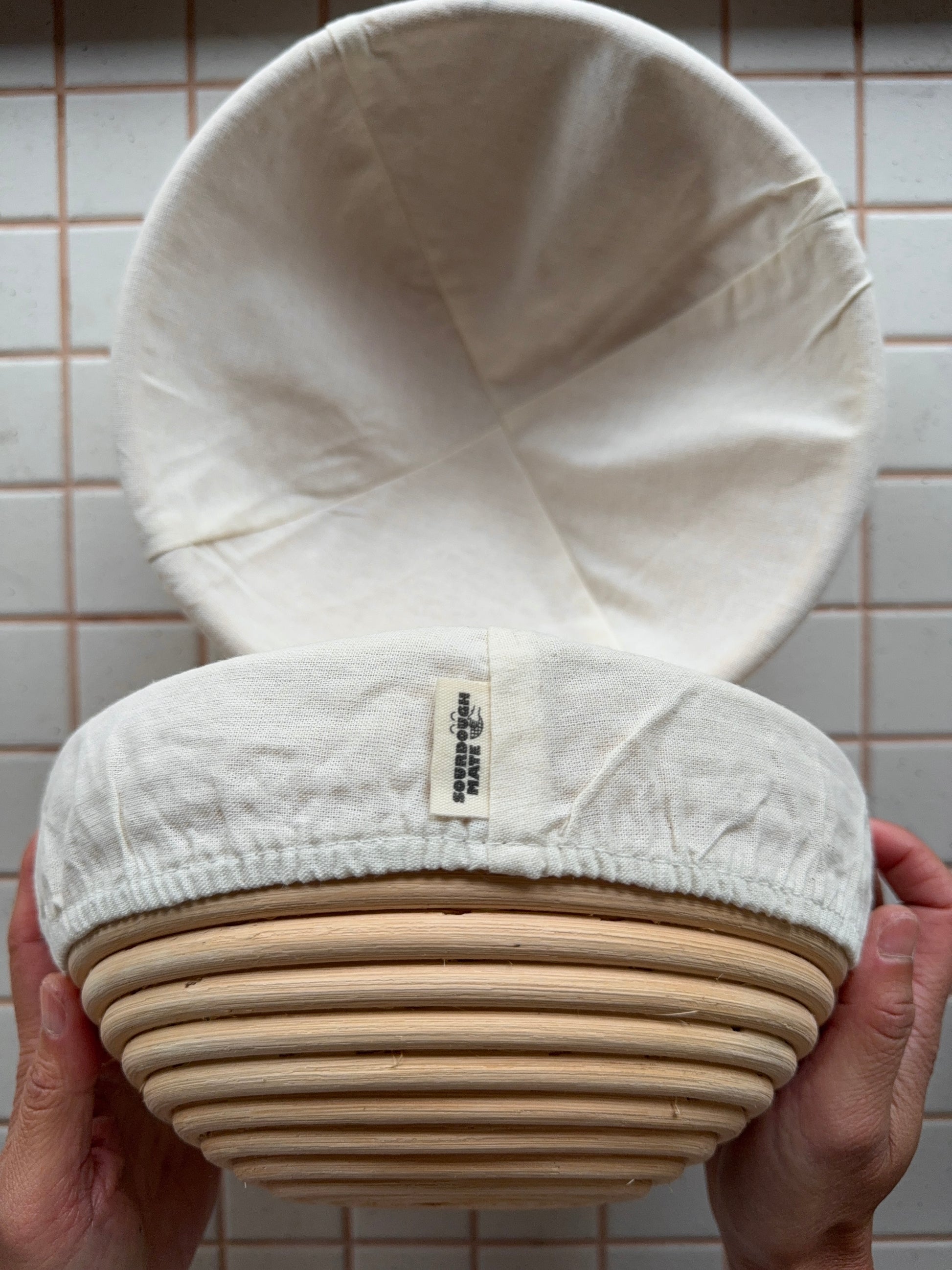 White fabric-covered bread proofing basket held by hands against a tiled wall background