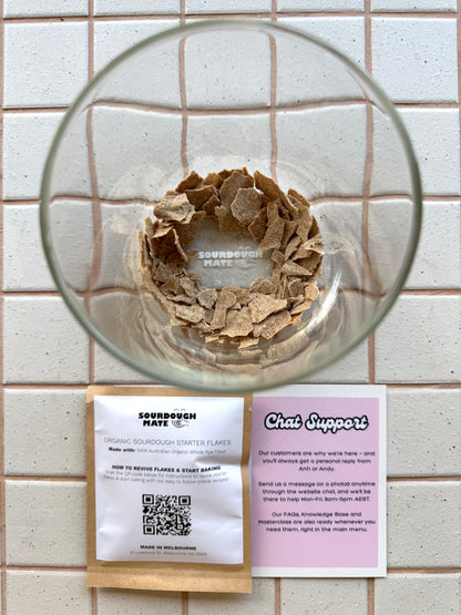 A clear glass bowl containing a brown flour-like starter culture, photographed from above on a white tiled surface. Below the bowl are two printed cards: one white titled 'Sourdough Mates Organic Sourdough Starter Flakes' and one pink titled 'Chat Support'.