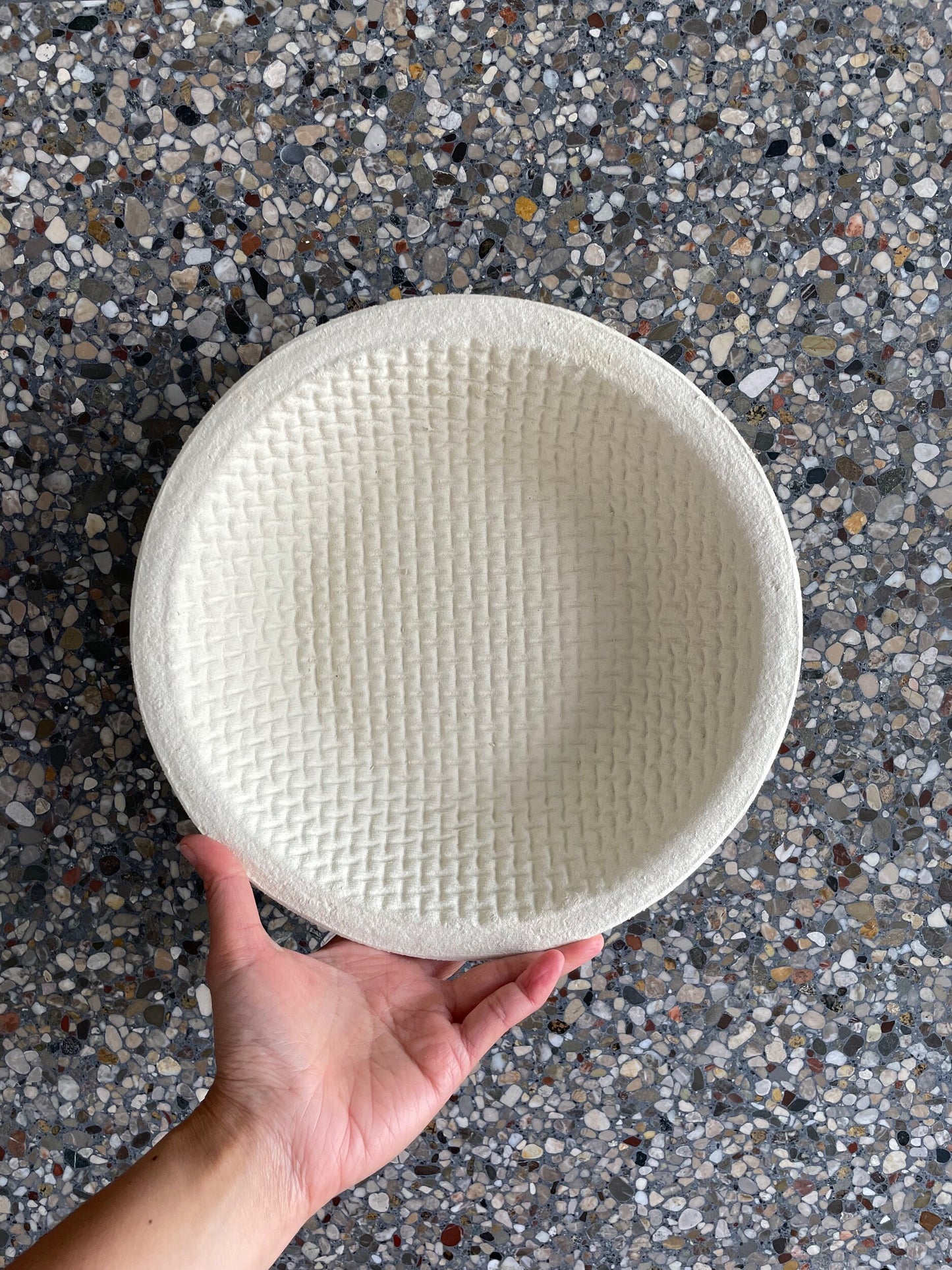 A hand holding a round beige wood pulp banneton with a waffle pattern texture, with pebble stone background
